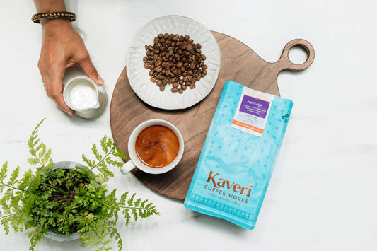 Kaveri Coffee on a wooden circle plank with loose coffee beans, a cup of coffee, and a hand holding a cup of milk to pour